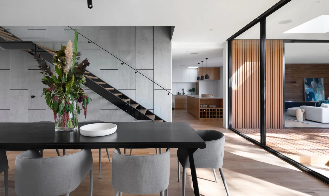Modern dining area with a black table, gray chairs, glass vase with green and purple plants, staircase with glass railing, and adjoining kitchen and living space with wood accents.